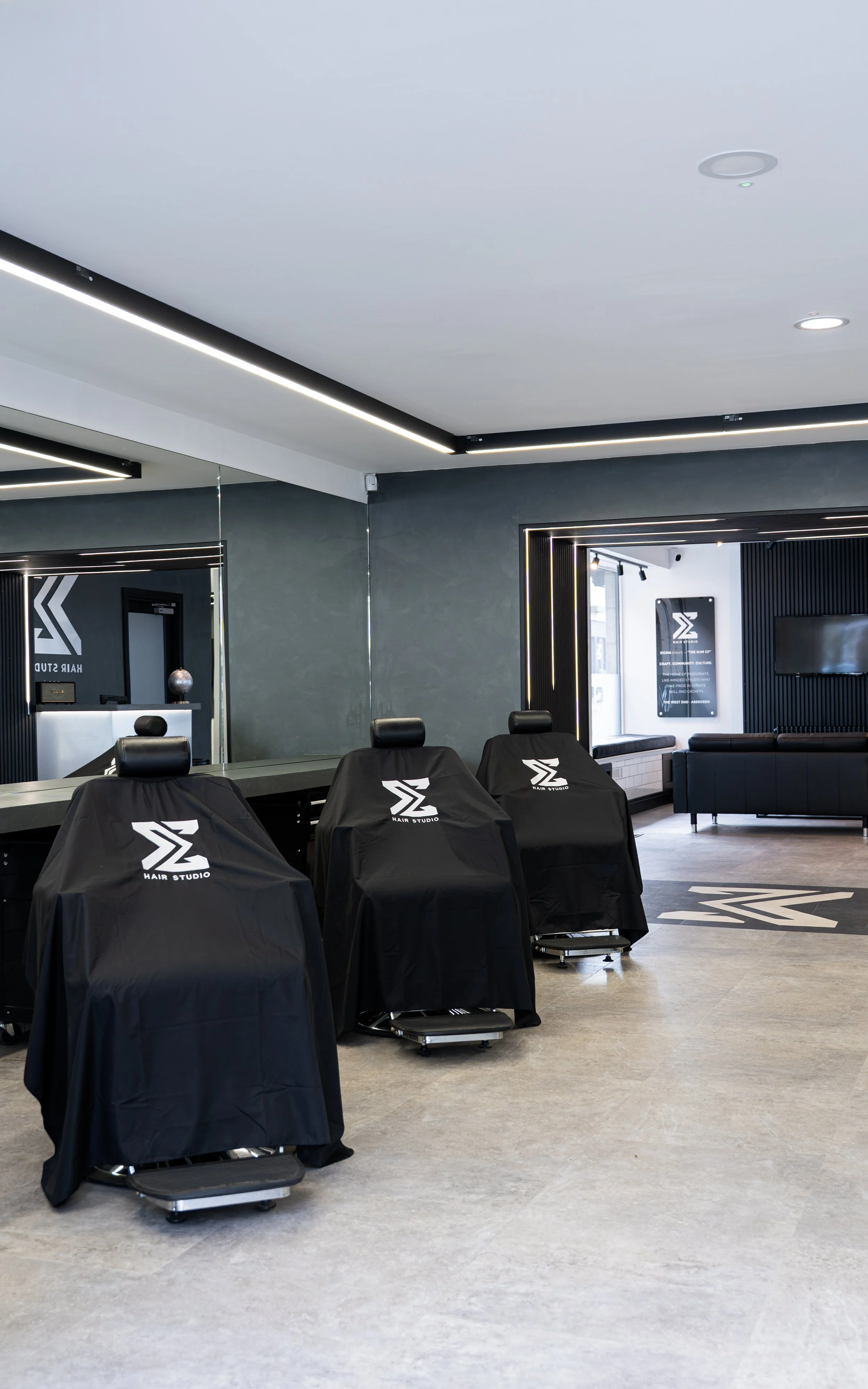 Interior of a modern hair salon with black chairs covered in black salon capes branded with 'Hair Studio' logo, a large mirror, and minimalist decor including black and white accents.