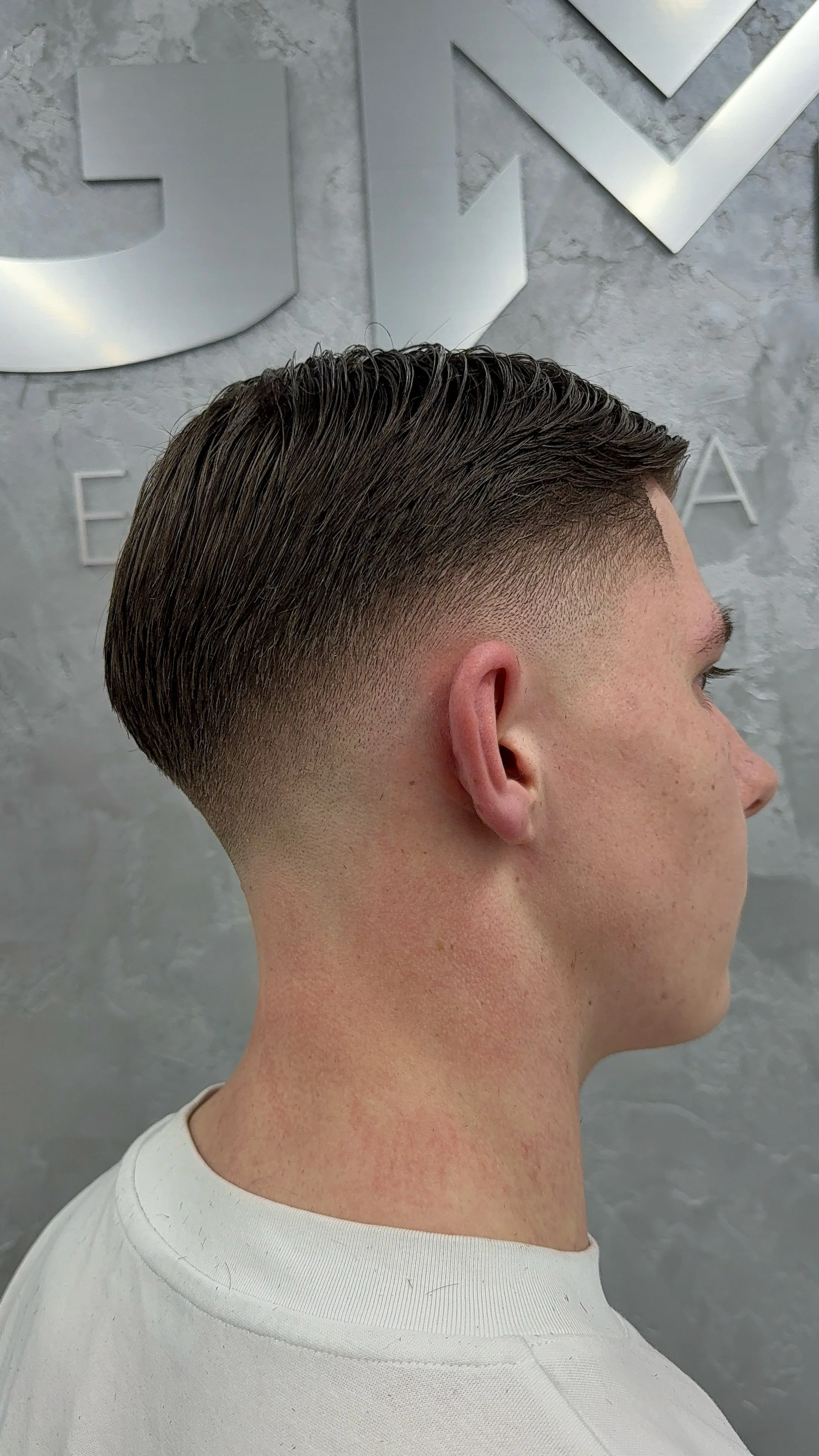 Side view of a man with a modern fade haircut, dark brown hair, and a clean-shaven face in front of a gray textured wall with large metallic letter decor.