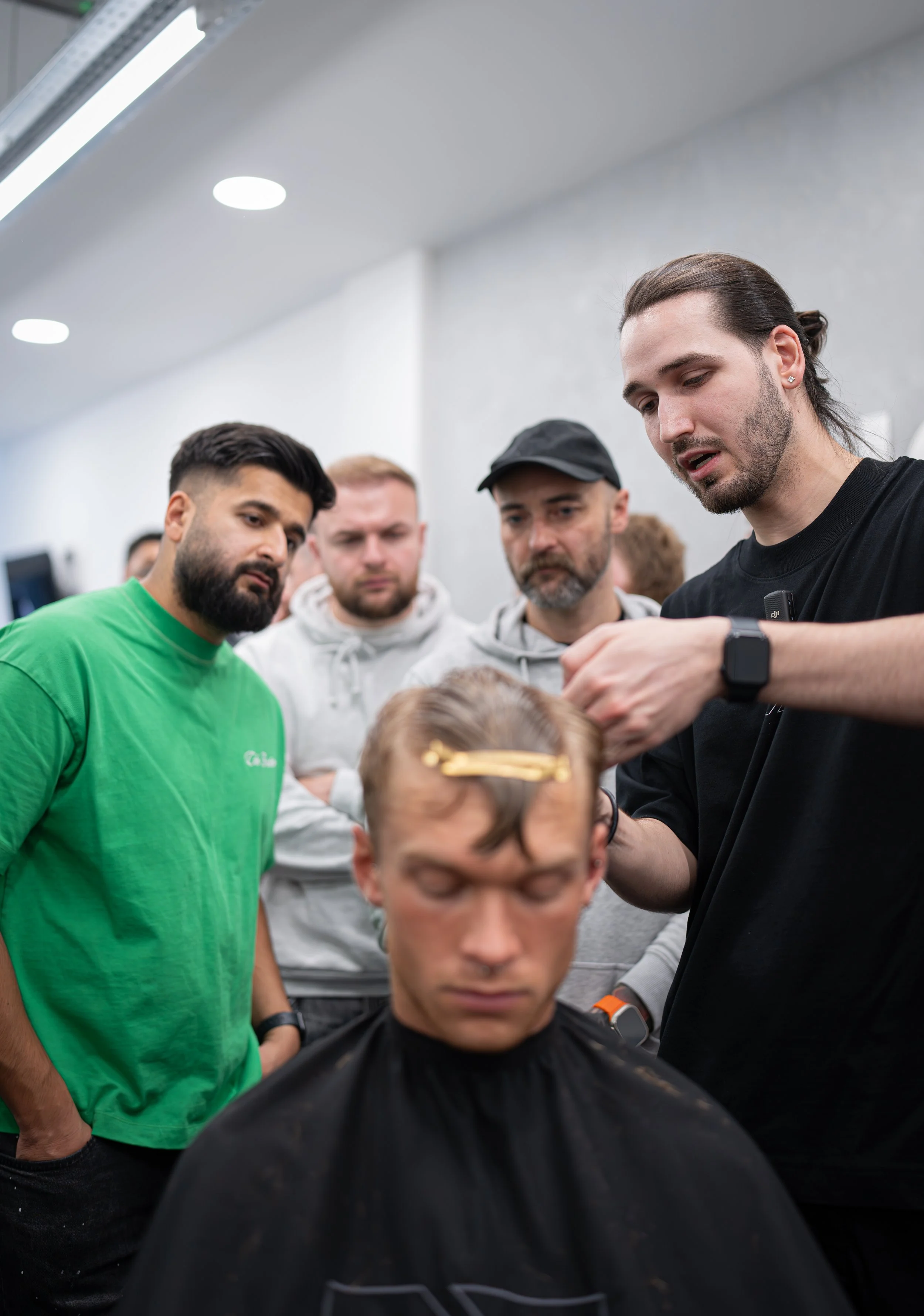 Barber giving haircut to a young man while four men observe in a barbershop.