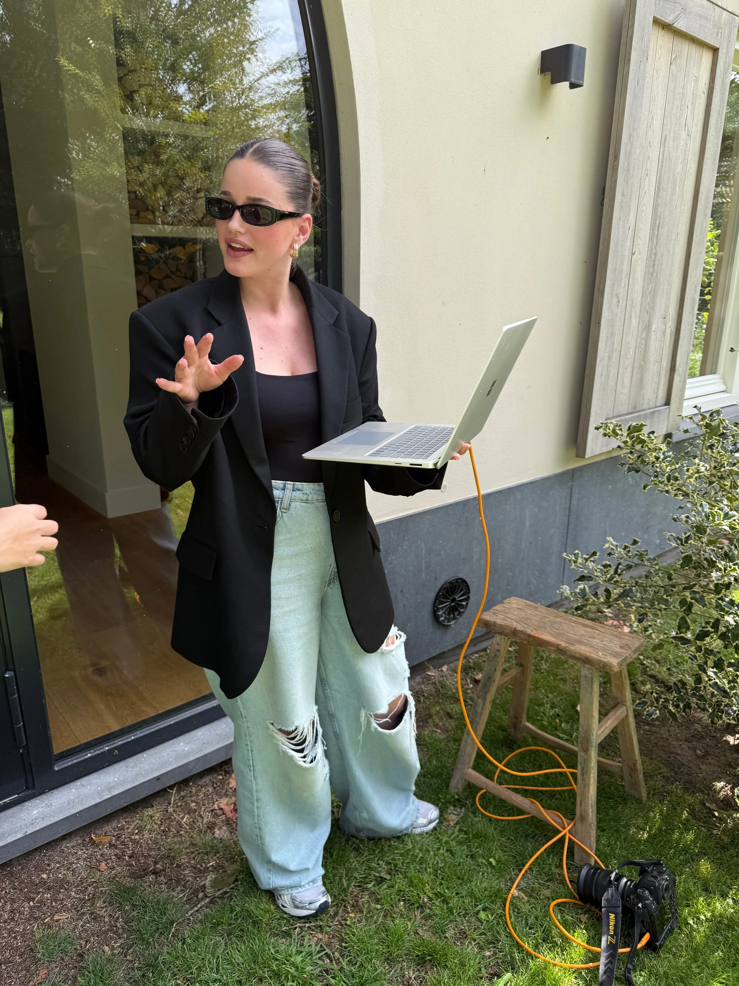A woman wearing black sunglasses, a black blazer, and ripped jeans standing outdoors near a house, holding a silver laptop connected to a camera on a small wooden stool via an orange cable, while talking to someone off-camera.
