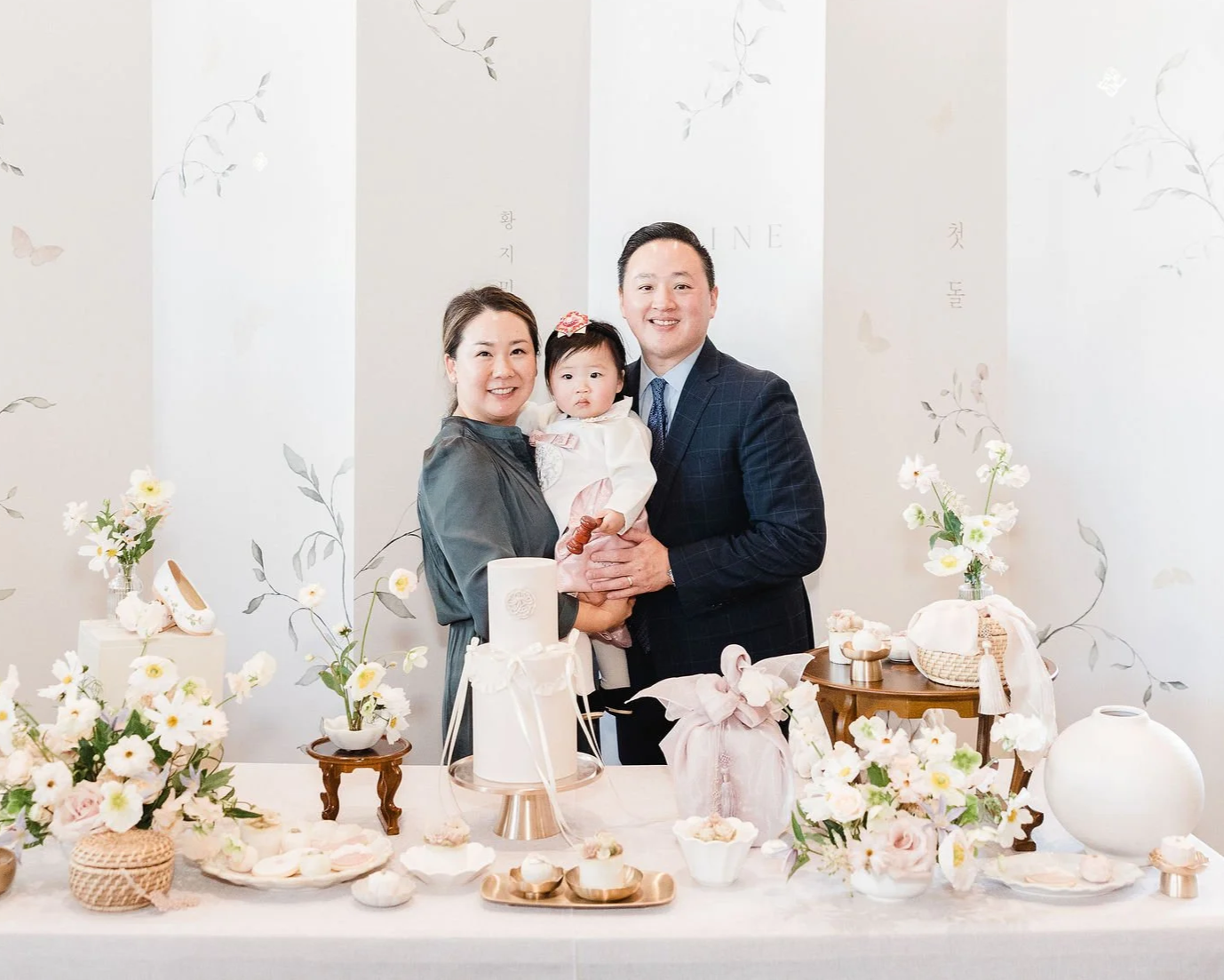 Family celebrating a birthday or special occasion with decorated table and cake, including two adults and a young child with floral decor in the background.