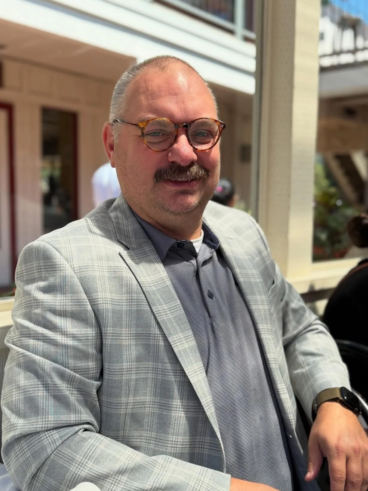 A man with glasses and a mustache, wearing a gray plaid blazer and a patterned shirt, sitting at a table indoors with a window in the background.