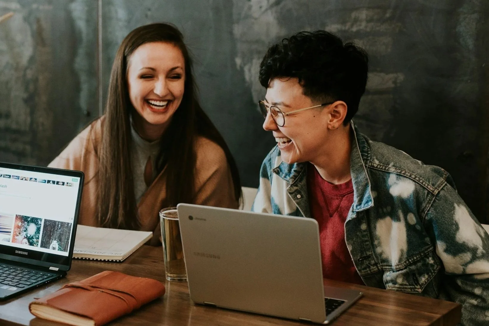 Two women sitting at a table, smiling and laughing with each other. There are laptops, a notebook, a wallet, and a glass of drink on the table.