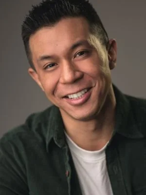A young man with short black hair smiling, wearing a dark green shirt over a white t-shirt, against a gray background.