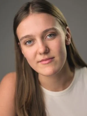 Close-up of a young woman with long brown hair, light complexion, and blue eyes, looking directly at the camera against a gray background.