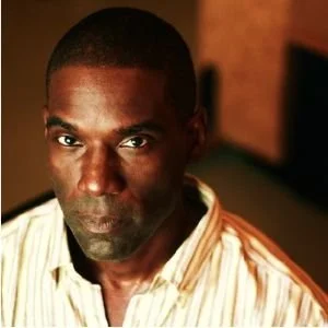 Close-up portrait of a man with dark skin and short hair, wearing a light-colored striped shirt, in an indoor setting with warm lighting.