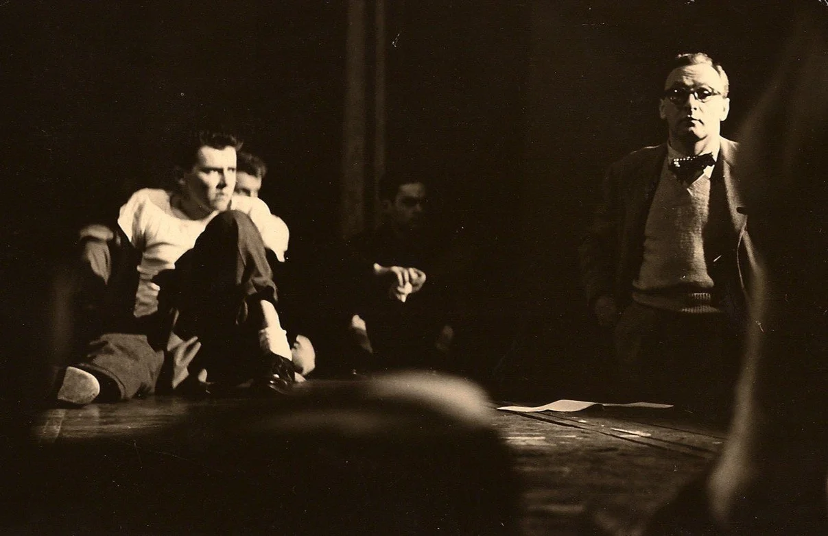 Black and white photo of a man standing and three seated men in a dimly lit room, with a dark background.