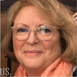 Close-up of a smiling woman with glasses and shoulder-length blonde hair, wearing a pink scarf.