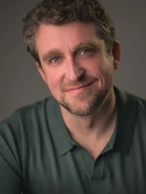 A man with curly brown hair and a beard smiling at the camera, wearing a dark green shirt against a gray background.