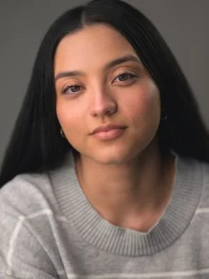 Portrait of a young woman with long black hair, wearing a gray sweater, looking directly at the camera against a neutral background.