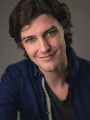 A young man with dark, wavy hair and fair skin, smiling slightly, wearing a dark blue shirt and a necklace, against a gray background.