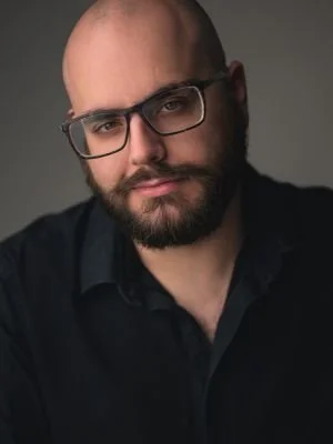 A man with a bald head, beard, and glasses posing against a neutral background.