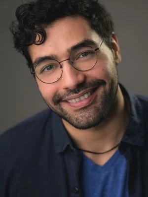 Portrait of a young man with curly dark hair, glasses, and a beard, smiling at the camera with a gray background.