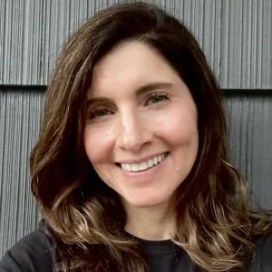 A woman with long brown hair smiling in front of a gray wooden wall.