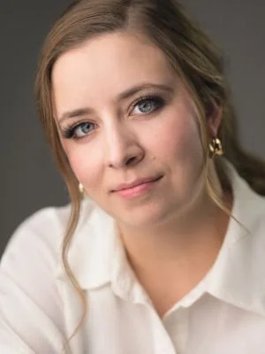 Close-up of a young woman with light brown hair, blue eyes, and gold hoop earrings, wearing a white shirt against a gray background.