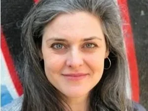 A woman with gray hair and blue eyes smiling, wearing a small hoop earring, with a red and black background.