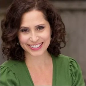Close-up of a woman with curly brown hair, smiling, wearing a green top, with a blurred background.
