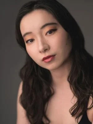 A young woman with long dark hair and natural makeup, looking at the camera against a dark gray background.