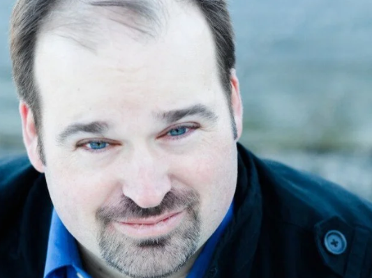 Close-up of a man with short brown hair, a beard, and blue eyes smiling outdoors.