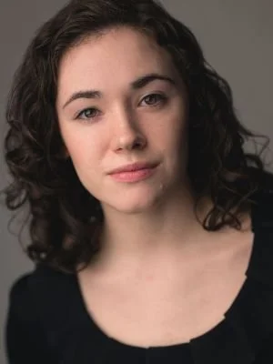 A young woman with dark, curly hair and fair skin, wearing a black top, looking at the camera against a plain gray background.
