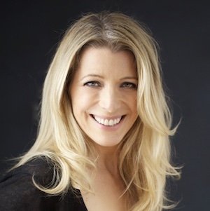 A woman with long blonde hair smiling at the camera, dark background