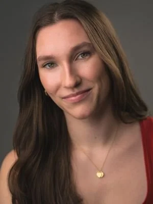 A young woman with long brown hair, wearing a red top and a gold necklace with a heart pendant, smiling at the camera.