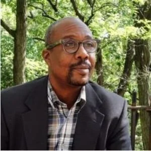 A middle-aged man with glasses, wearing a checkered shirt and blazer, sitting outdoors in a wooded area.