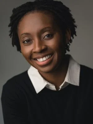 A smiling woman with natural hair, wearing a white collared shirt and black sweater, against a neutral background.