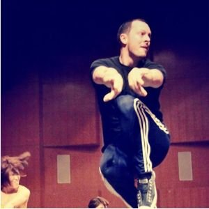 A person balancing on one foot with arms extended forward in a dance or performance setting, with a dark background.