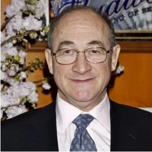 A middle-aged man with glasses, wearing a suit and tie, standing in front of floral decorations and a wooden background.
