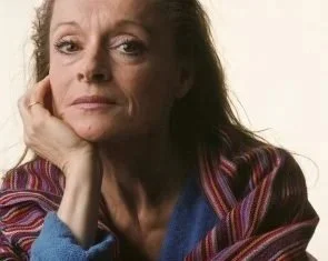 A woman with long hair and a serious expression, resting her chin on her hand, wearing a colorful striped jacket.