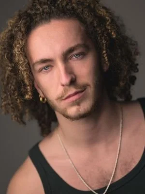Close-up portrait of a young man with curly hair and blue eyes, wearing a black tank top and a necklace