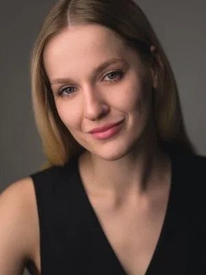 A young woman with blonde hair wearing a black sleeveless top, smiling gently at the camera.