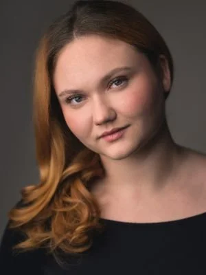 A young woman with light skin and reddish-brown hair styled in loose waves, wearing a black top, looking at the camera with a neutral expression.