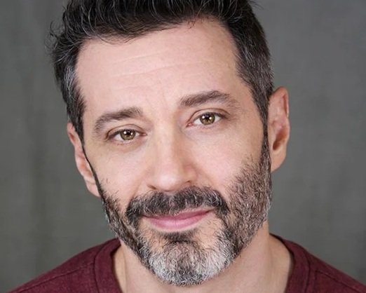Close-up portrait of a middle-aged man with dark hair, a beard, and a gentle smile, wearing a maroon shirt against a gray background.