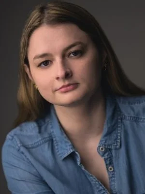 Young woman with long brown hair wearing a blue denim shirt, looking at the camera with a neutral expression.