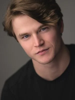 Close-up portrait of a young man with light brown hair, blue eyes, and fair skin, wearing a black shirt.
