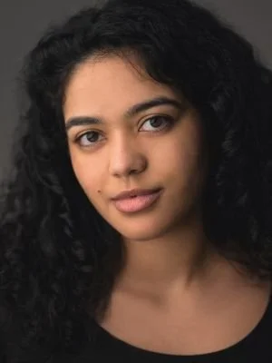 A close-up portrait of a young woman with dark, curly hair and brown eyes, wearing a black top against a dark background.