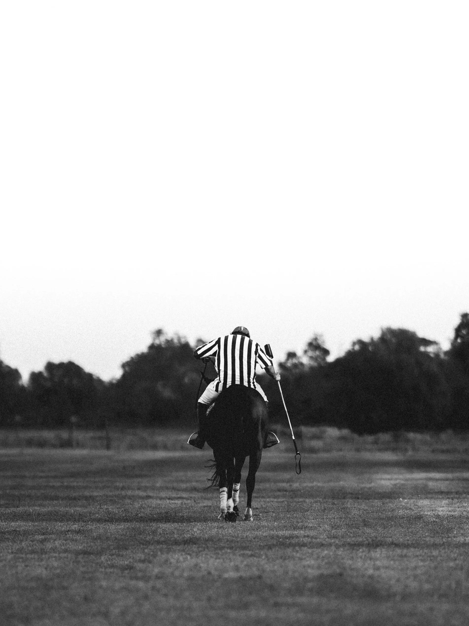 A jockey riding a horse on a race track, dressed in a striped shirt, holding a riding crop, with trees in the background, in black and white.
