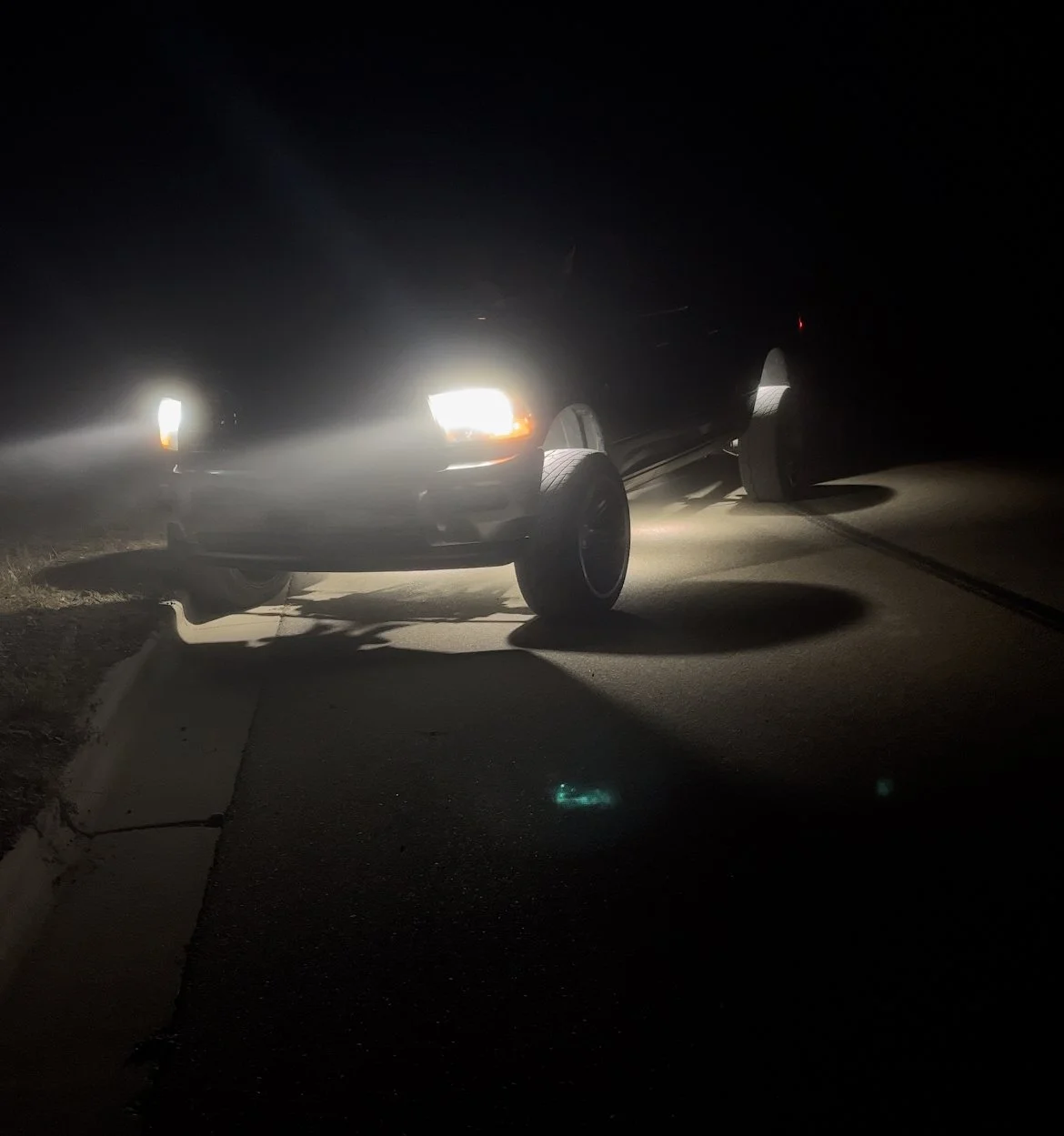 A black truck is parked on the side of a dark road at night with its headlights on, casting bright light and shadows on the pavement.
