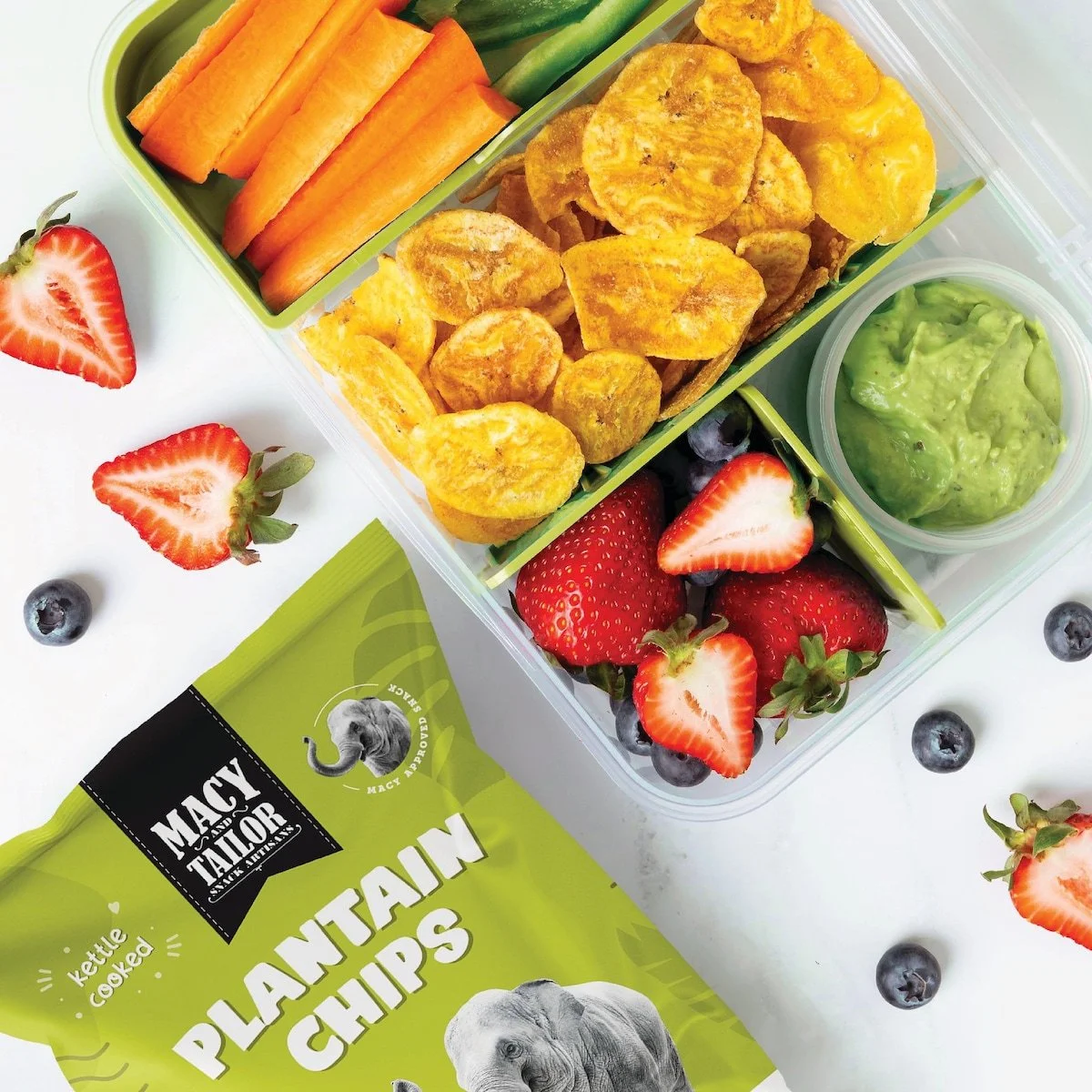 A divided lunchbox with fresh strawberries, blueberries, sliced carrots, cucumber sticks, popcorn, and guacamole, with scattered berries and strawberries around on a white surface and a bag of Maitai Mountain plantain chips.