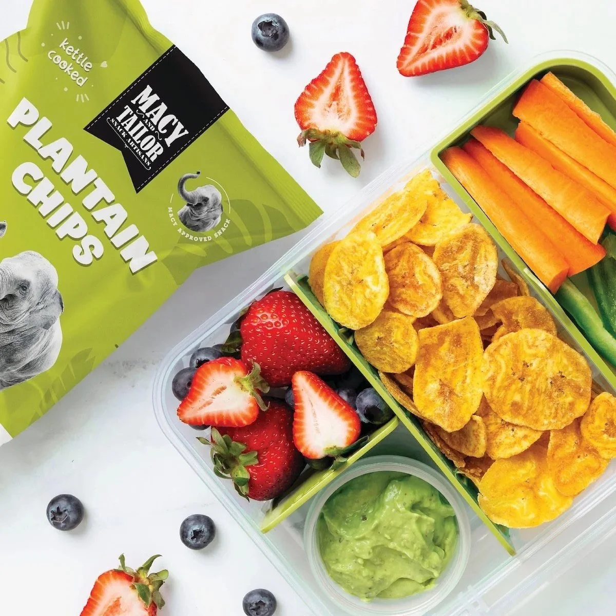 A healthy lunchbox containing strawberries, blueberries, tortilla chips, sliced carrots, cucumber sticks, guacamole, and chicken nuggets, with a green packaging bag labeled 'Plantain Chips' by Macy Taylor.