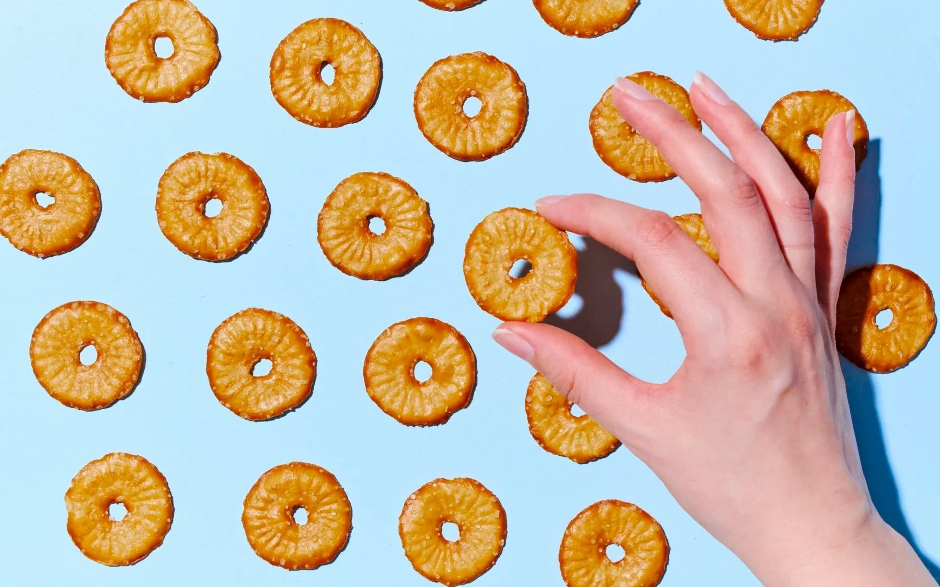 A hand reaches out to pick up a circular, orange, glazed pastry on a pale blue background, surrounded by similar pastries arranged in a grid pattern.