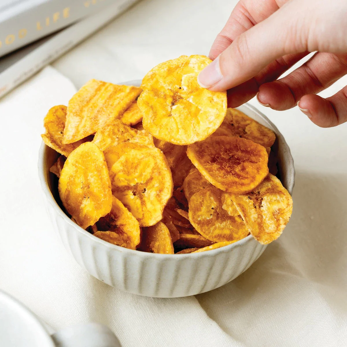 Hand reaching to pick a banana chip from a white bowl filled with banana chips.