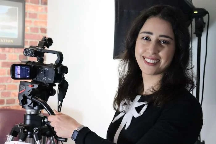 A woman with dark hair stands next to a professional video camera on a tripod, smiling at the camera.