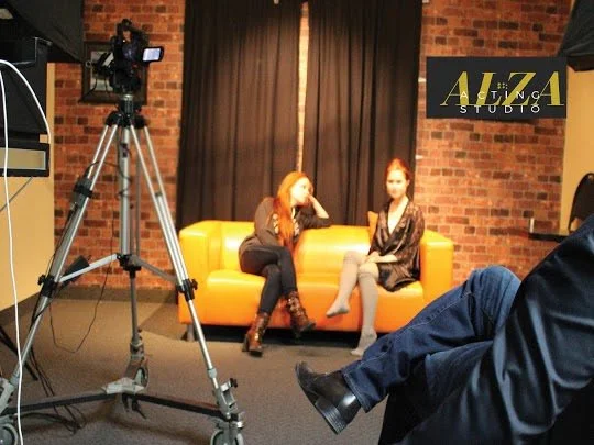 Two women sitting on an orange couch in a recording studio with a camera on a tripod, a person in the foreground, and a sign that reads Aliza Studio on the wall.
