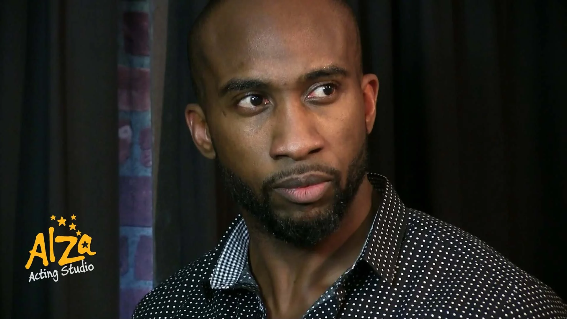A man with a beard looking to the side, wearing a black shirt with white dots, with a background of black curtains and a small section of patterned fabric on the left. The logo in the bottom left corner says 'AliZa Acting Studio' in yellow and white text.