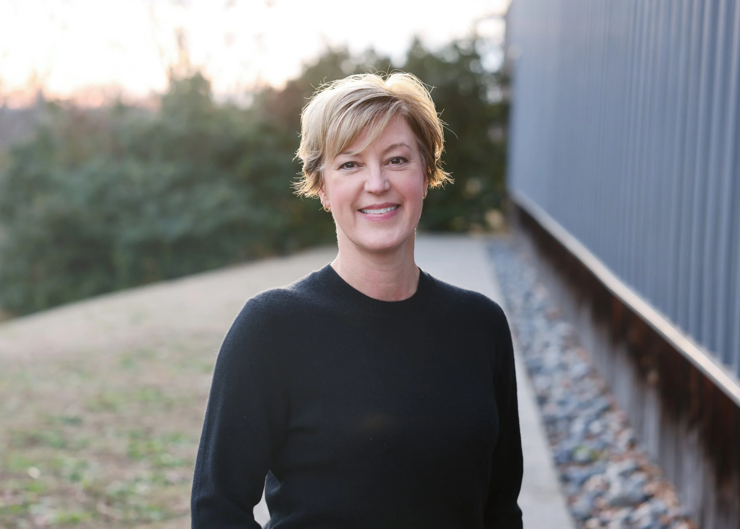 A woman with short blonde hair smiling outdoors near a blue building with trees in the background.