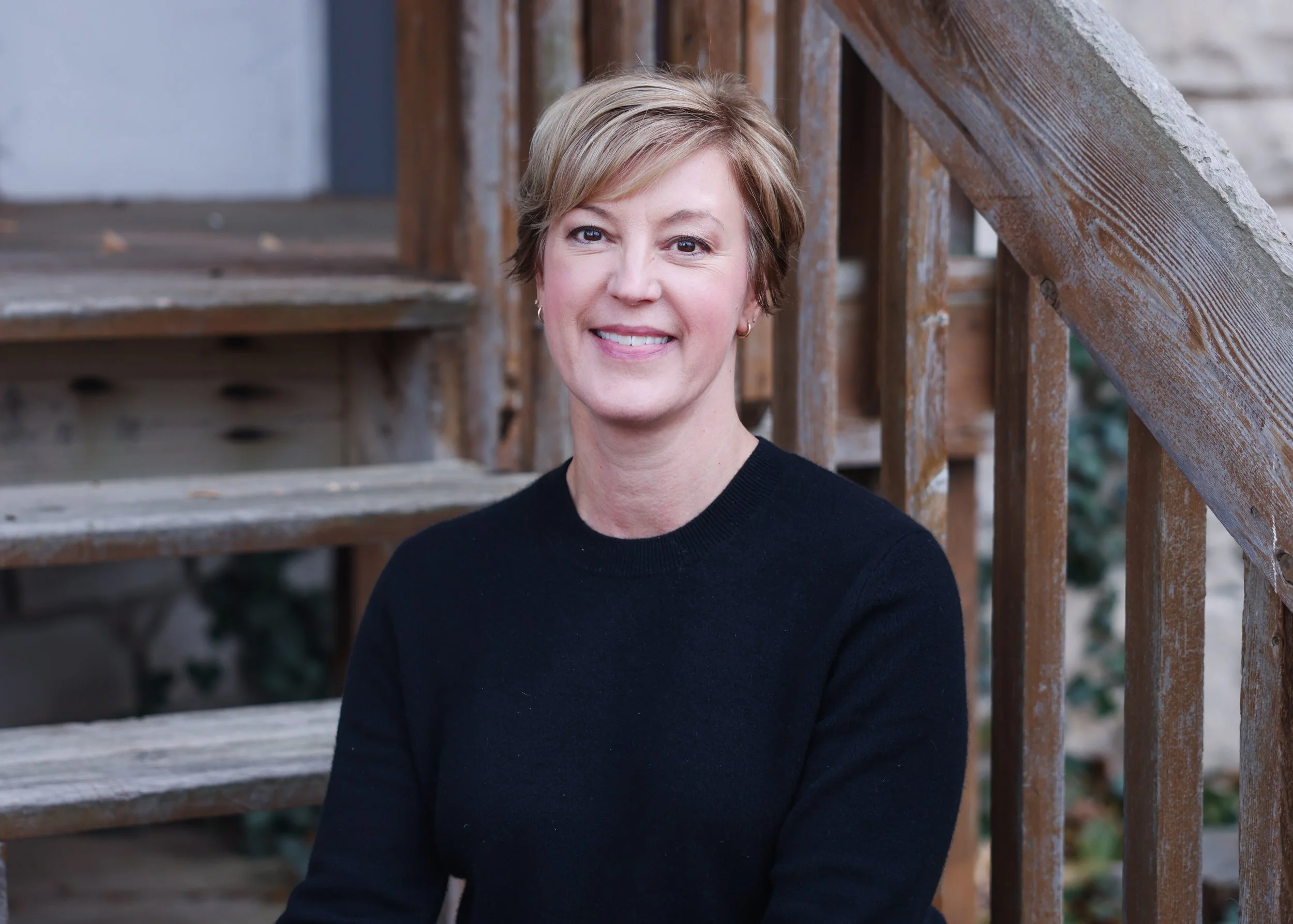 A woman with short blond hair and earrings, smiling, standing in front of a wooden staircase railing outdoors.