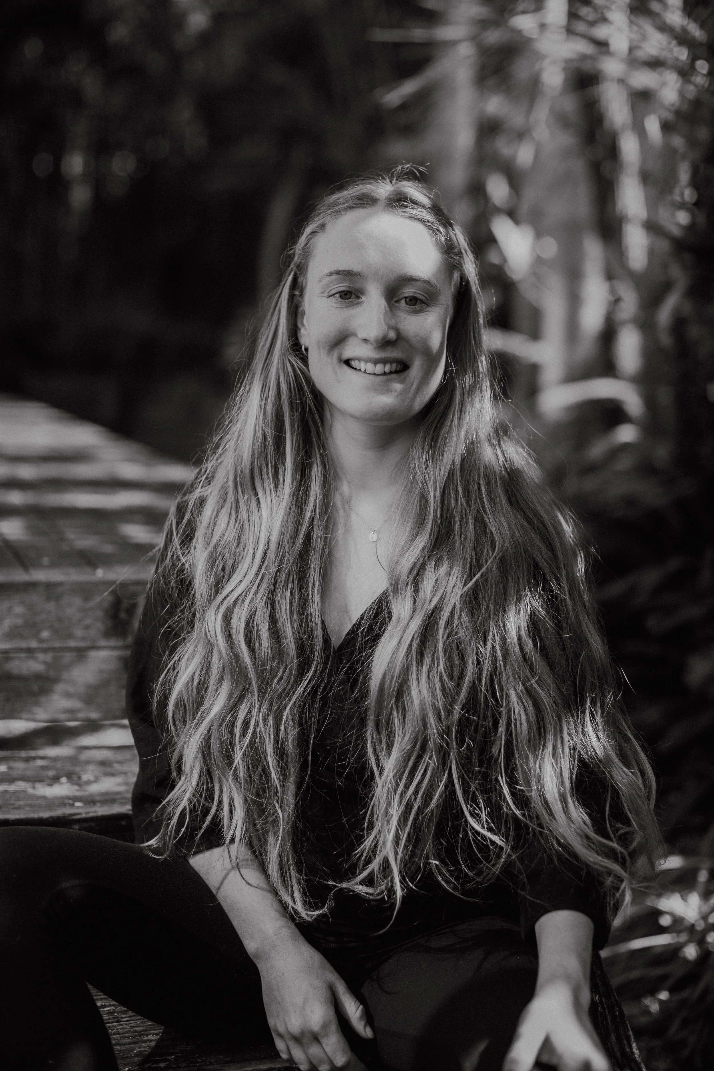 Black and white portrait of a smiling woman with long wavy hair sitting outdoors on a wooden bench surrounded by blurred foliage.